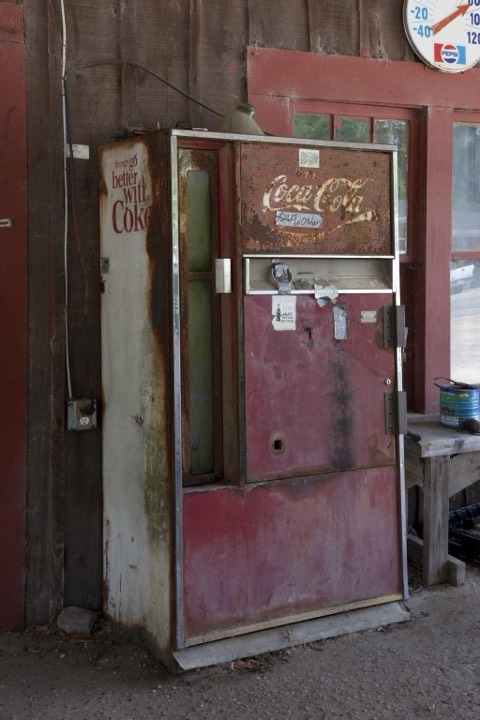 A weathered antique Coca-Cola vending machine with a chrome coin mechanism, under a porch